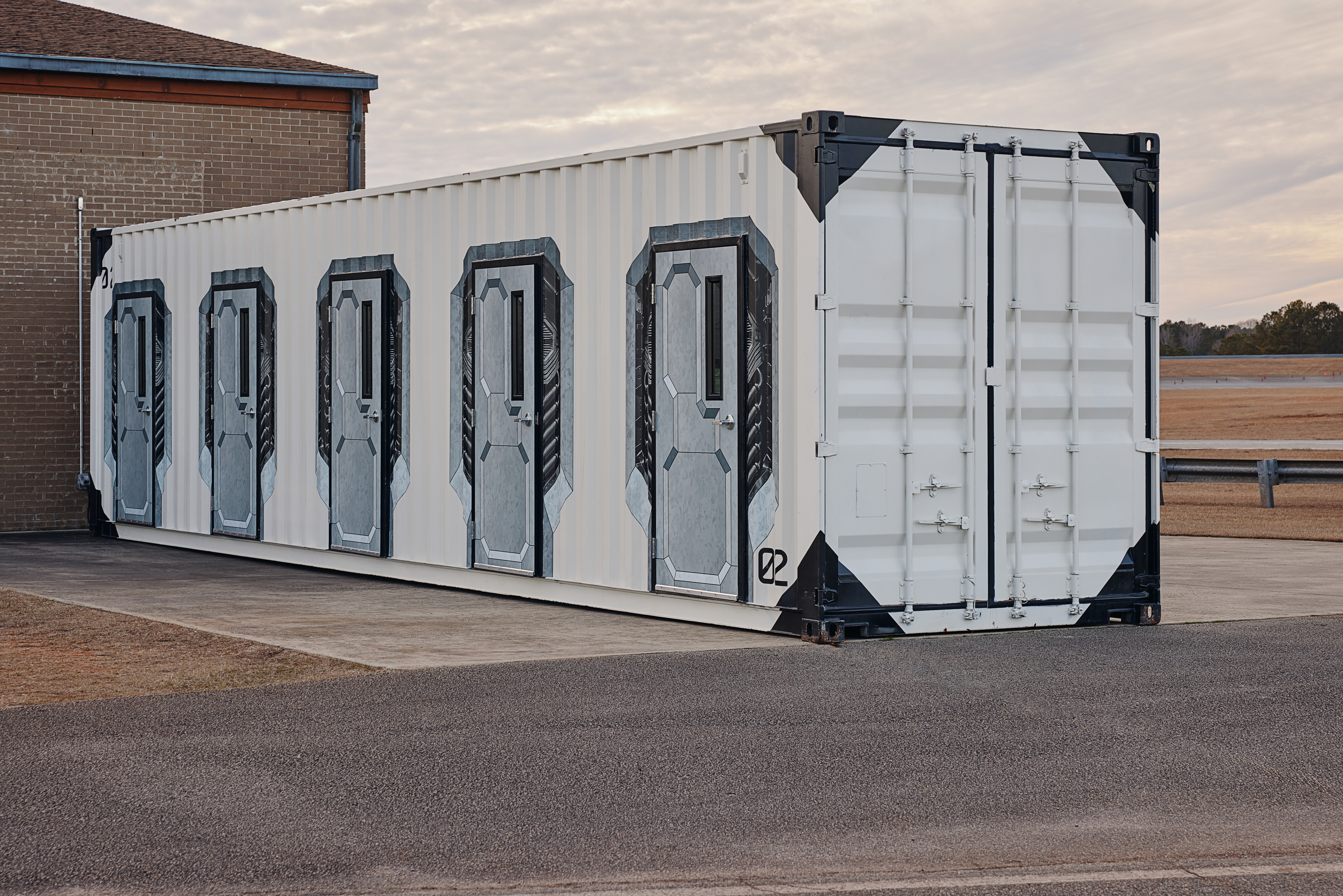 An Axon Training Pod positioned on pavement outside of a building. The container is white with six identical metal doors along one side, each framed in a decorative, futuristic design.
